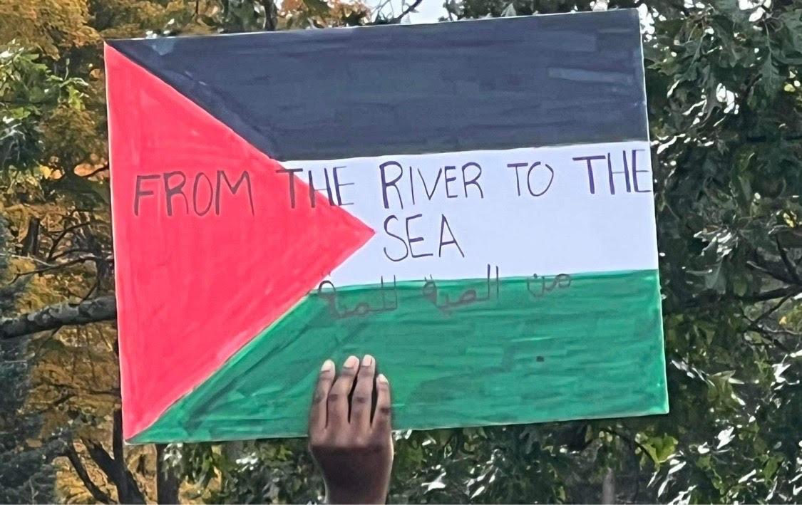 Student holding sign of Palestinian flag labeled "From the river to the sea"